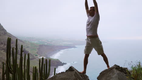 Al-Practicar-Yoga-De-Saludo-Al-Sol-Con-Un-Telón-De-Fondo-Montañoso-Y-Vistas-Al-Mar,-Un-Joven-En-Forma-Abraza-La-Meditación-Y-El-Ejercicio,-Destacando-Un-Estilo-De-Vida-Deportivo-Y-Saludable