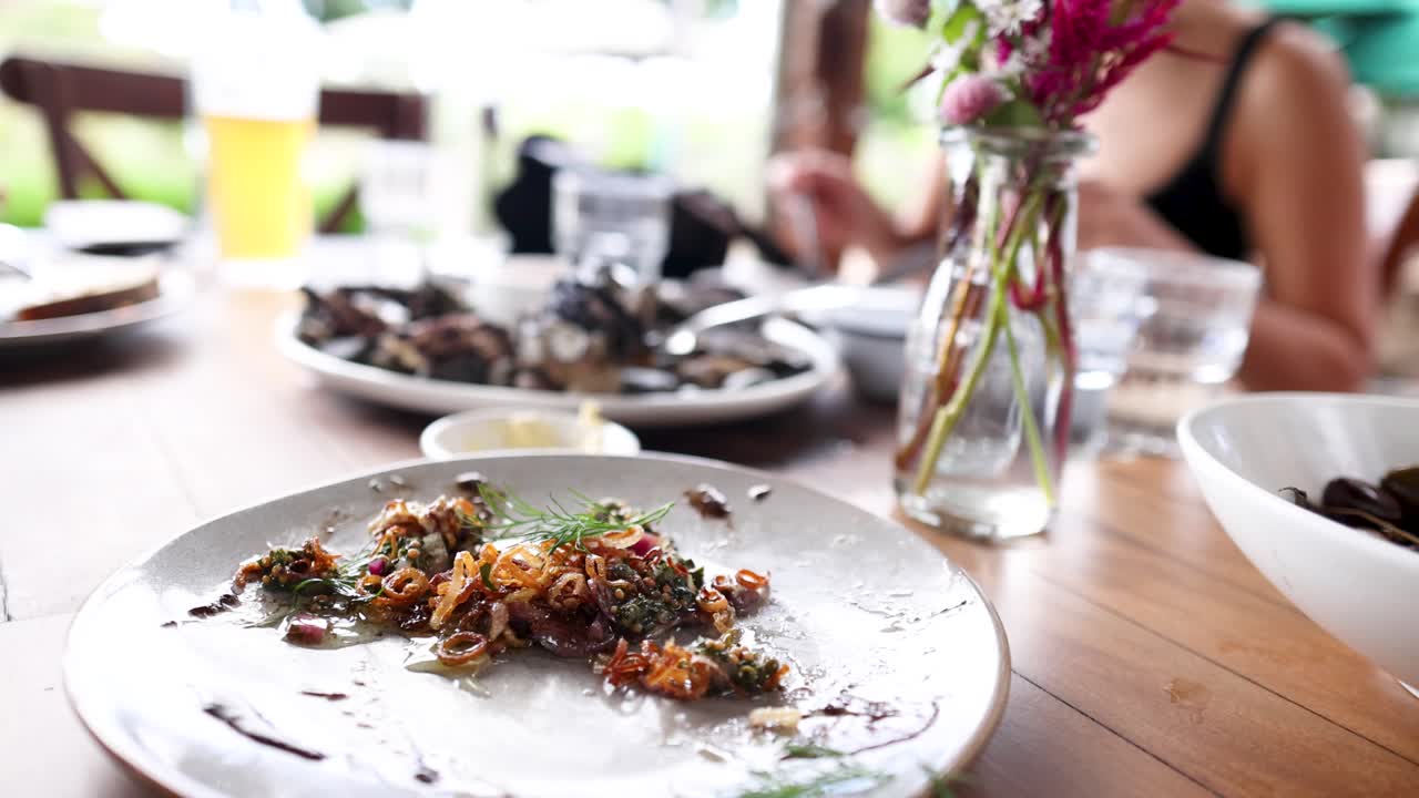 Diners enjoy a meal at a sunlit restaurant table, featuring vibrant dishes and a relaxed atmosphere