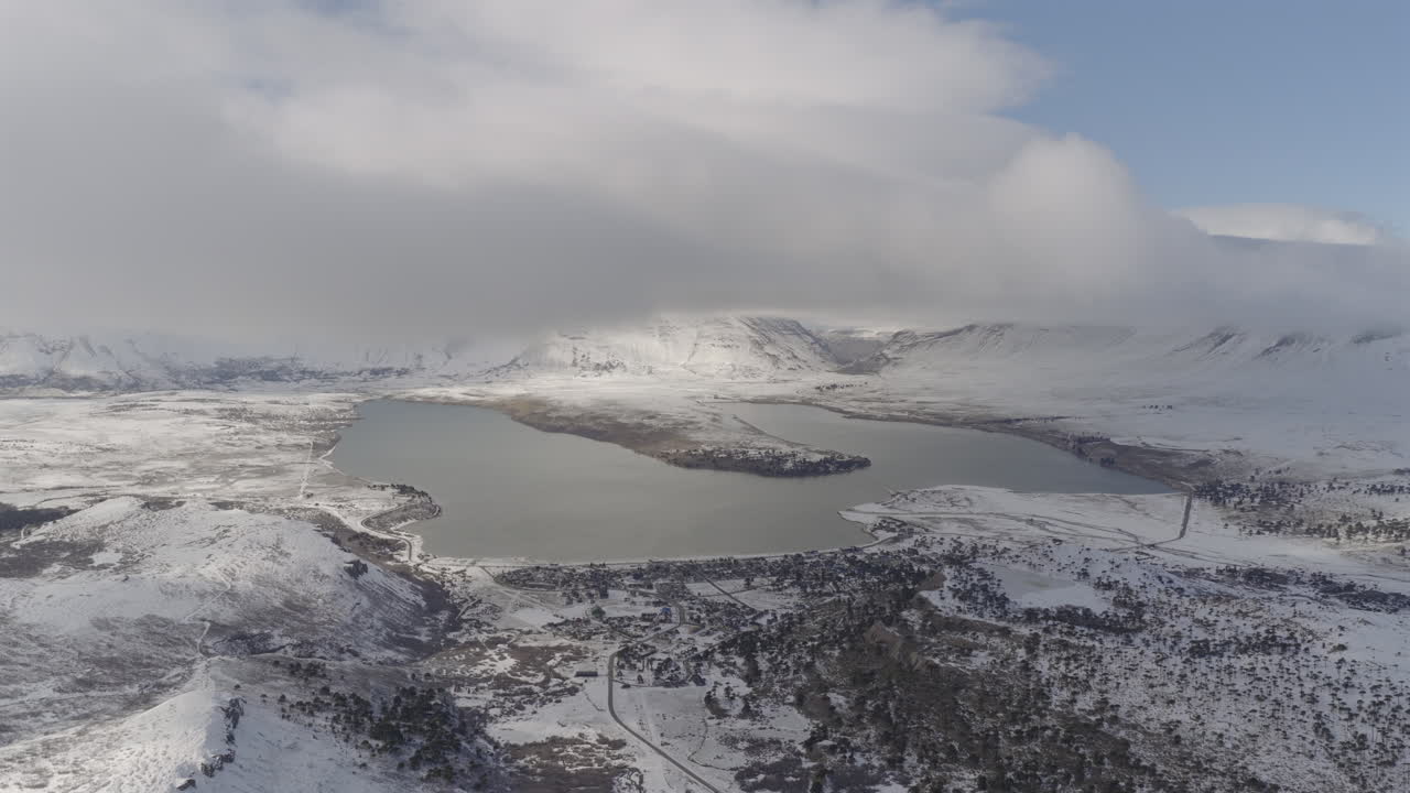 Lake Caviahue in the Caviahue-Copahue area, Neuquén Province, Argentina with snow-covered Andes mountains framing frozen lake in the foreground, stratus cloud layer drifts across, Static aerial view