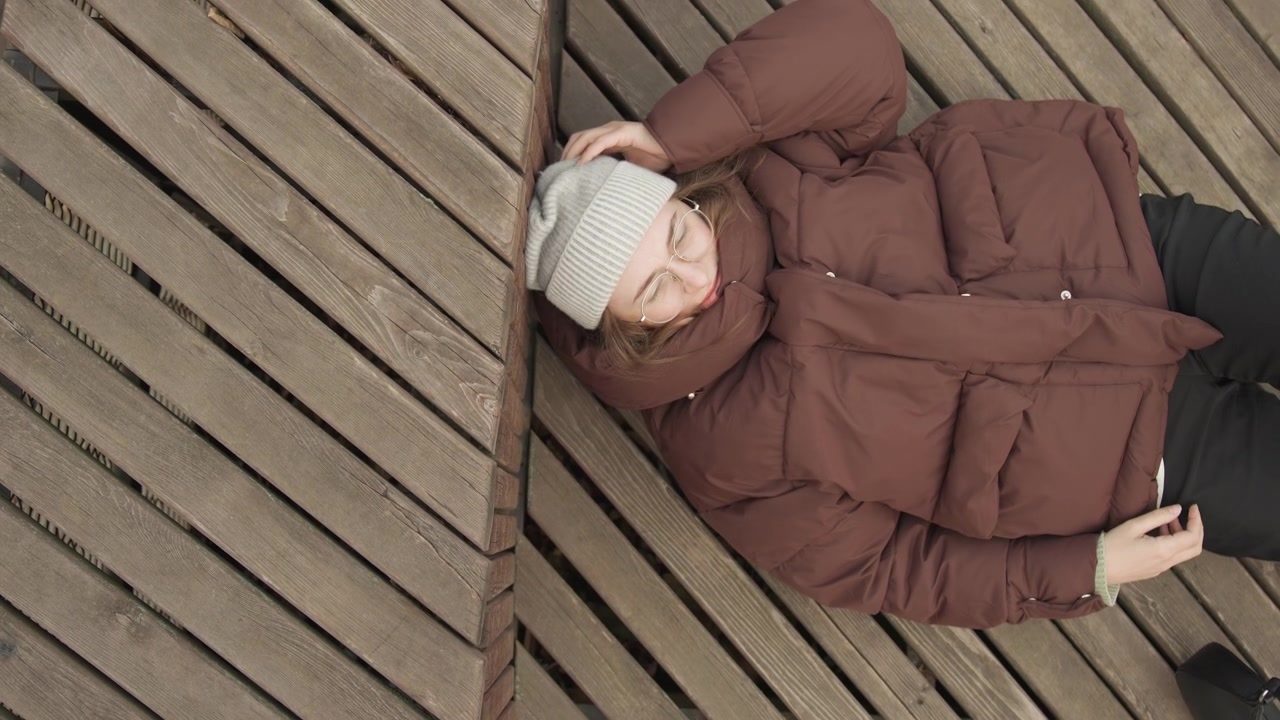 Relaxed woman in casual winter outfit. Woman enjoying cool weather on city boardwalk smiling happily. Relaxed woman dressed in winter clothes sitting on wooden path smiling on chilly day