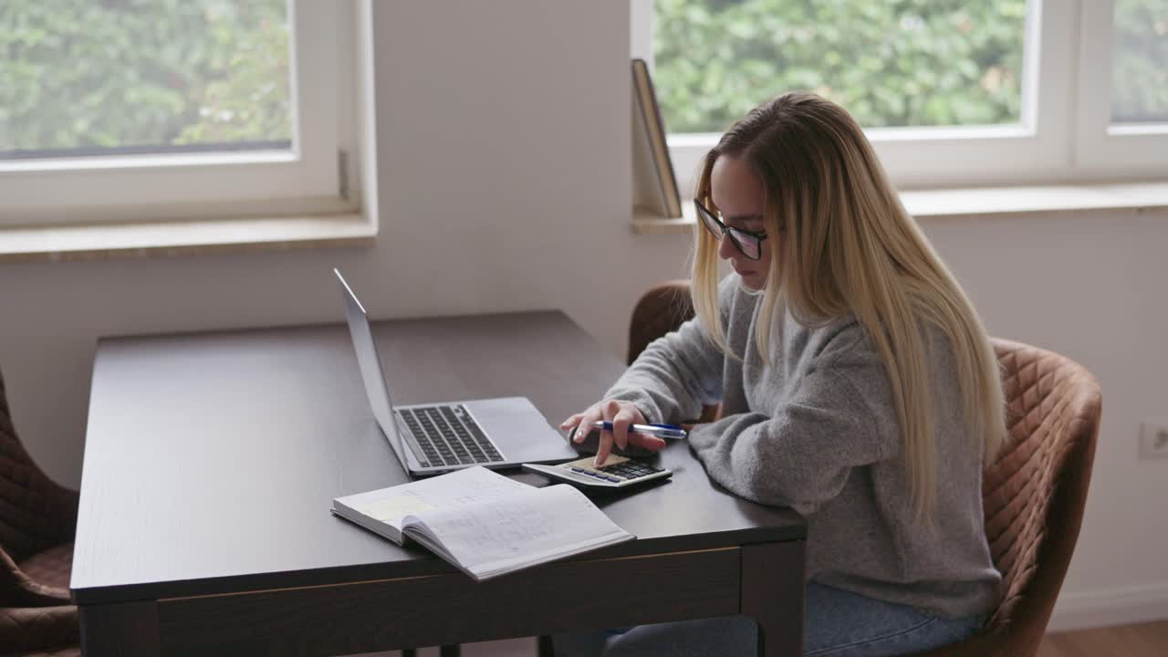 En ung kvinne med briller sitter ved et skrivebord hjemme, jobber på laptopen sin mens hun bruker en kalkulator.