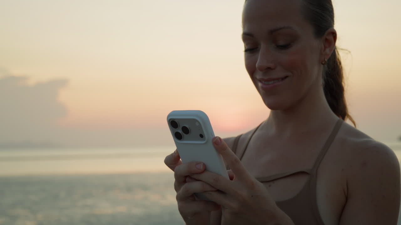 Giovane donna sorridente che usa lo smartphone al tramonto in riva al mare
