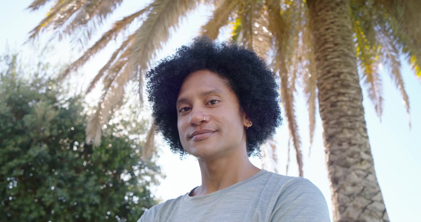 Afroameričan stojí venku u palmy, kamera se oddaluje, usmívá se v šedém tričku.