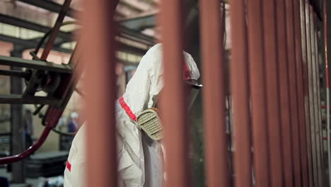 Painting process in workshop. A man in safety suit and respirator paints metal frame with a spray gun inside the small factory.