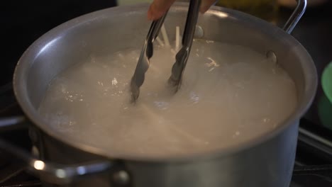 closeup shot of boiling noodles in an aluminium kitchen cooking utensil using a tong