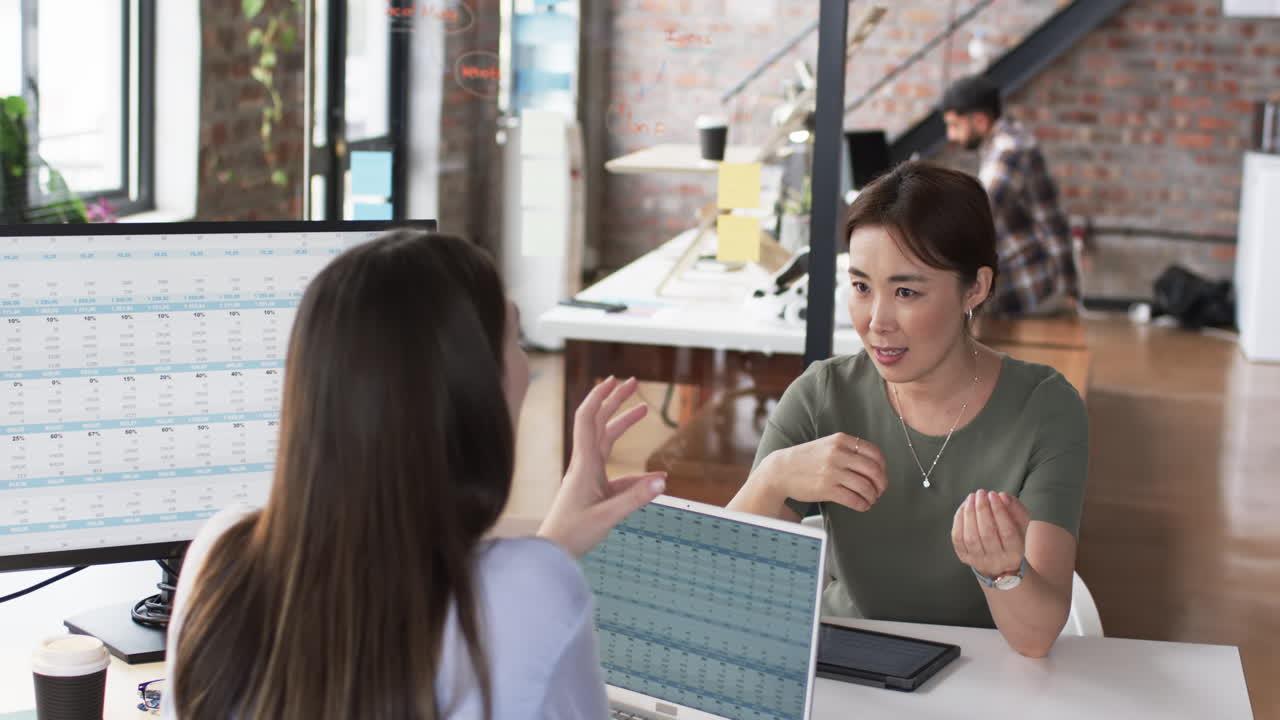 una mujer asiática con una camiseta verde discute negocios con una joven mujer caucásica a través de un monitor de computadora