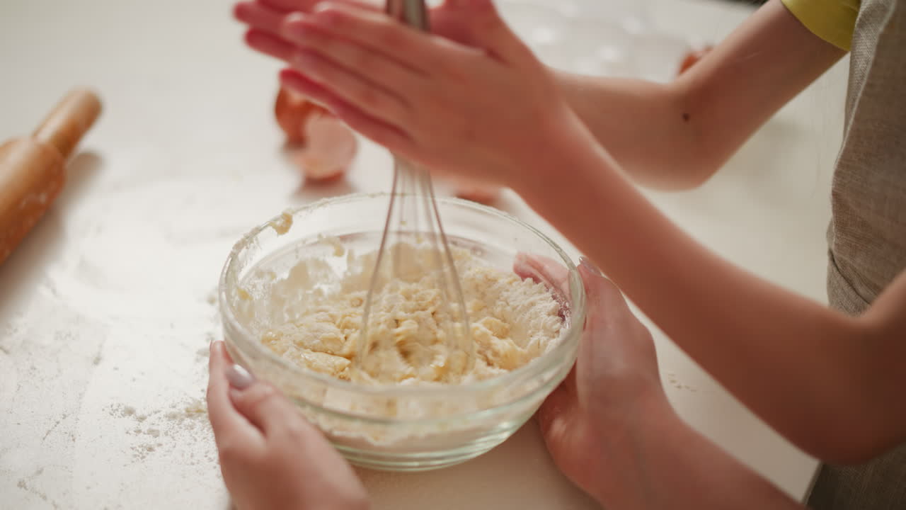 close up of baking activity showing one person whisking flour mixture while another holds glass bowl steady on flour-dusted white counter with broken eggshells and wooden rolling pin in background
