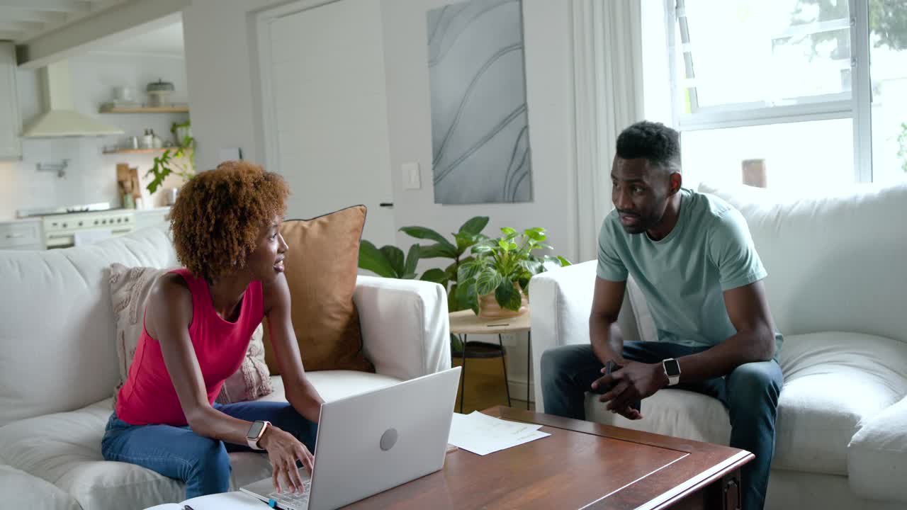 Diverse couple on sofa working on laptop when man entering showing smartphone, celebrating success