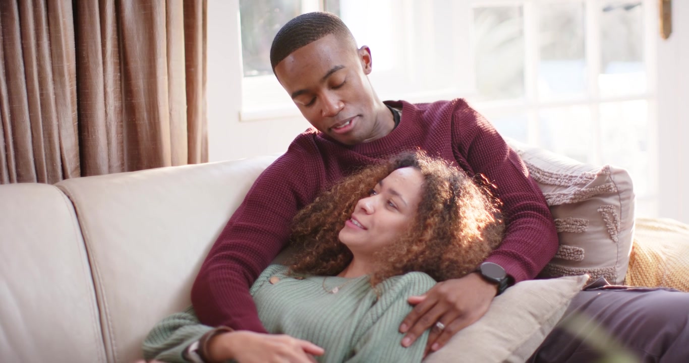 Couple reclining on light couch, woman resting head on lap while man adjusting pillow, comforting