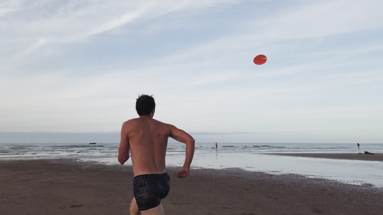 Back view of a shirtless man running toward the sea to catch an airborne orange frisbee as his friend looks on from afar, in Omaha Beach at low tide in dramatic slow motion