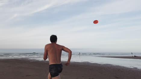 back view of a shirtless man running toward the sea to catch an airborne orange frisbee as his friend looks on from afar, in omaha beach at low tide in dramatic slow motion