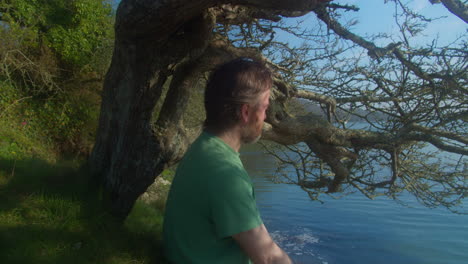 hombre barbudo de mediana edad disfruta del cálido sol de la mañana sentado al lado de un árbol mirando hacia el lago