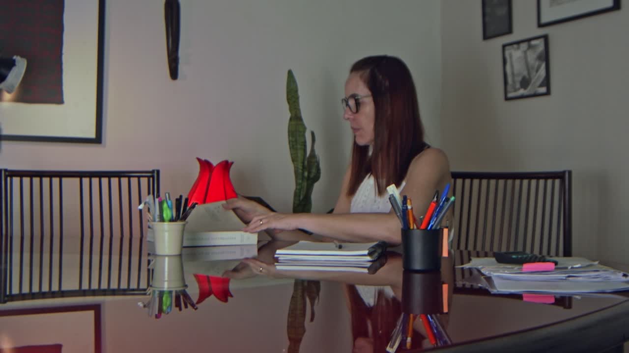 video estático de una mujer aplicada con anteojos sentada en su escritorio en casa tomando notas de un libro