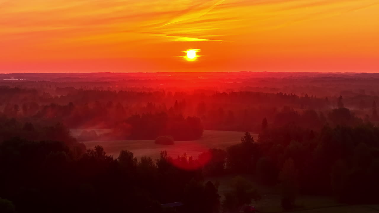 A brilliant sunrise casts warm orange and red hues over a misty forest landscape in Latvia, creating a tranquil and atmospheric scene