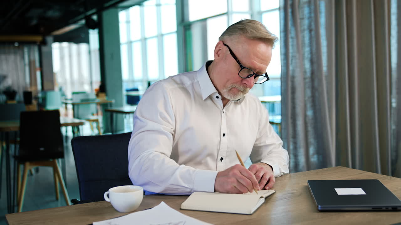 Bearded old man in white shirt writes in paper notebook. Man crosses the written and thinks for a moment.