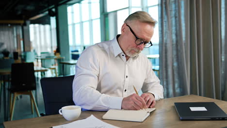 bearded old man in white shirt writes in paper notebook. man crosses the written and thinks for a moment.