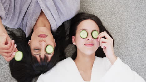 Happy-biracial-mother-and-adult-daughter-taking-cucumber-slices-from-eyes-and-laughing,-slow-motion