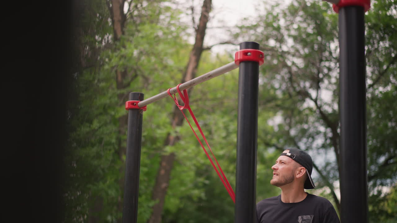 Man Securing Band Anchor Above Head On Outdoor Rig, Determined Trainer Ties Strap, Concentrated Expression, Vertical Metal Posts And Green Trees Frame Scene, Methodical Preparation For Workout