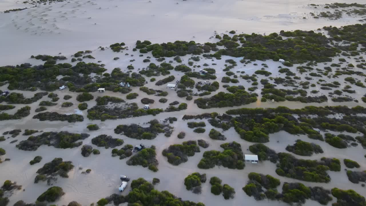 Panorâmica aérea de drone subindo sobre um parque de campismo à beira-mar com moinhos de vento ao nascer do sol.