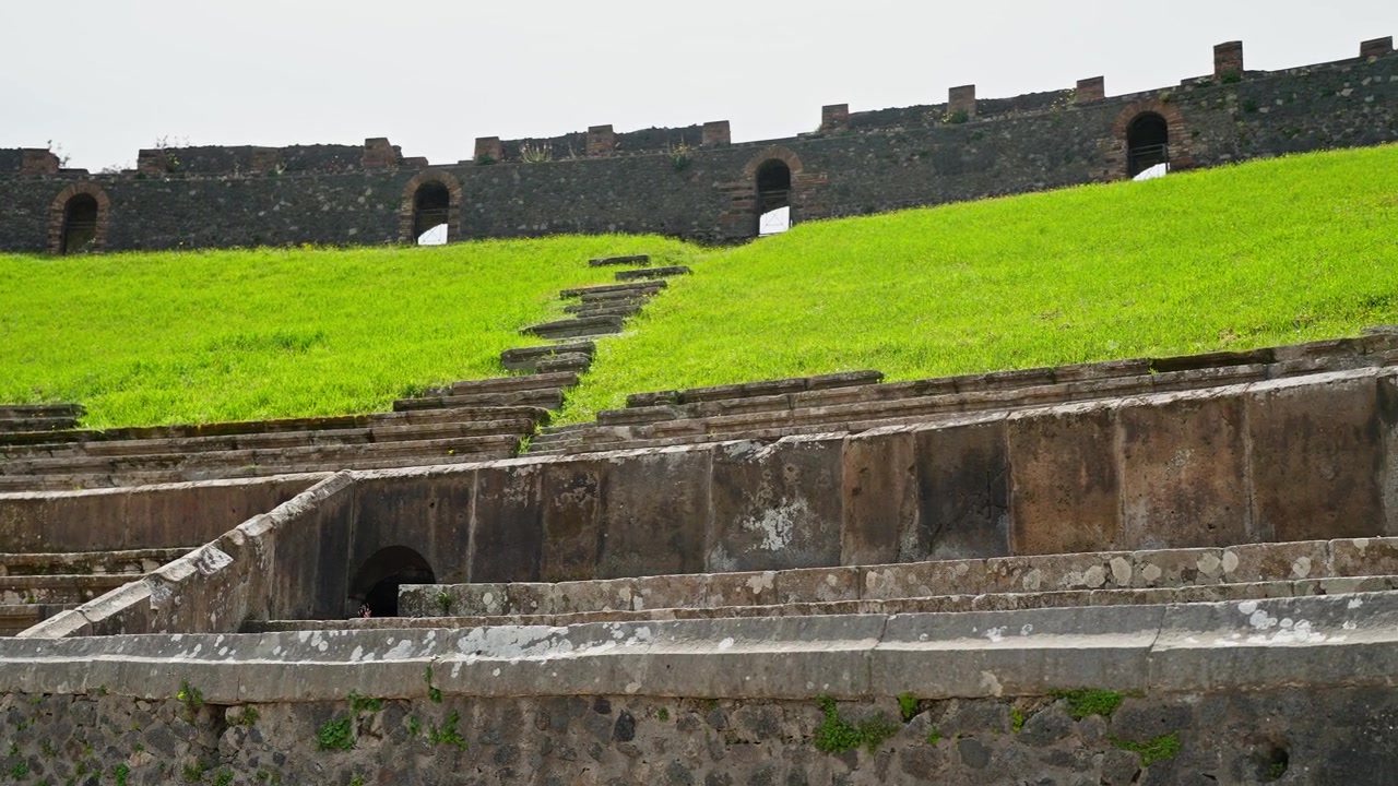 Interno dell'Anfiteatro di Pompei con prato verde dell'arena, antiche gradinate in pietra, cancelli ad arco e mura romane consumate dal tempo sotto un cielo coperto.