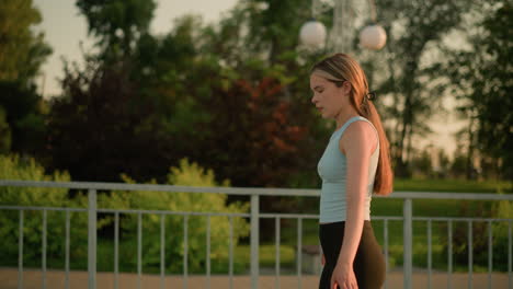 side view of lady roller skating near iron railing, gently swinging her hands, under warm evening sunlight, with lamp post, trees, and lush greenery in background