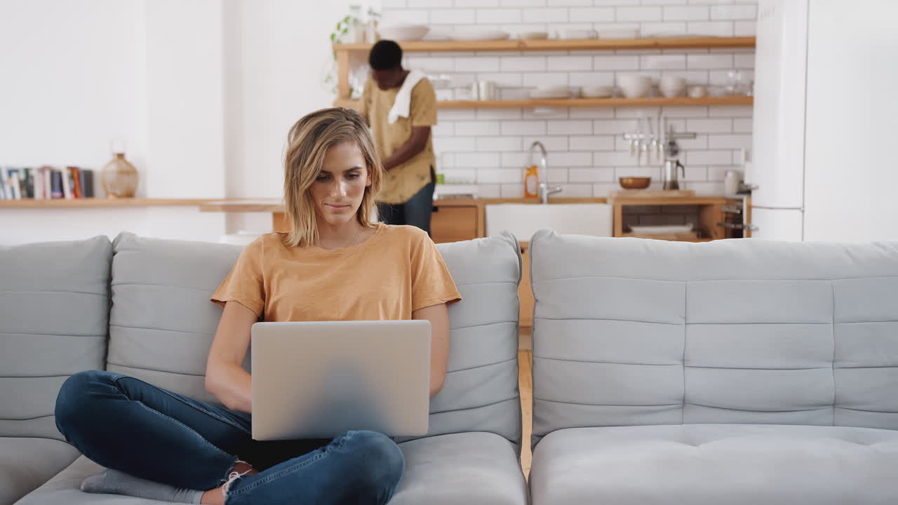 Woman On Sofa At Home Using Laptop Computer With Man In Kitchen Behind