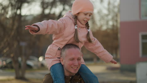 cheerful father and daughter moment with the girl in a pink cap and jacket, arms spread wide, sitting on his shoulders