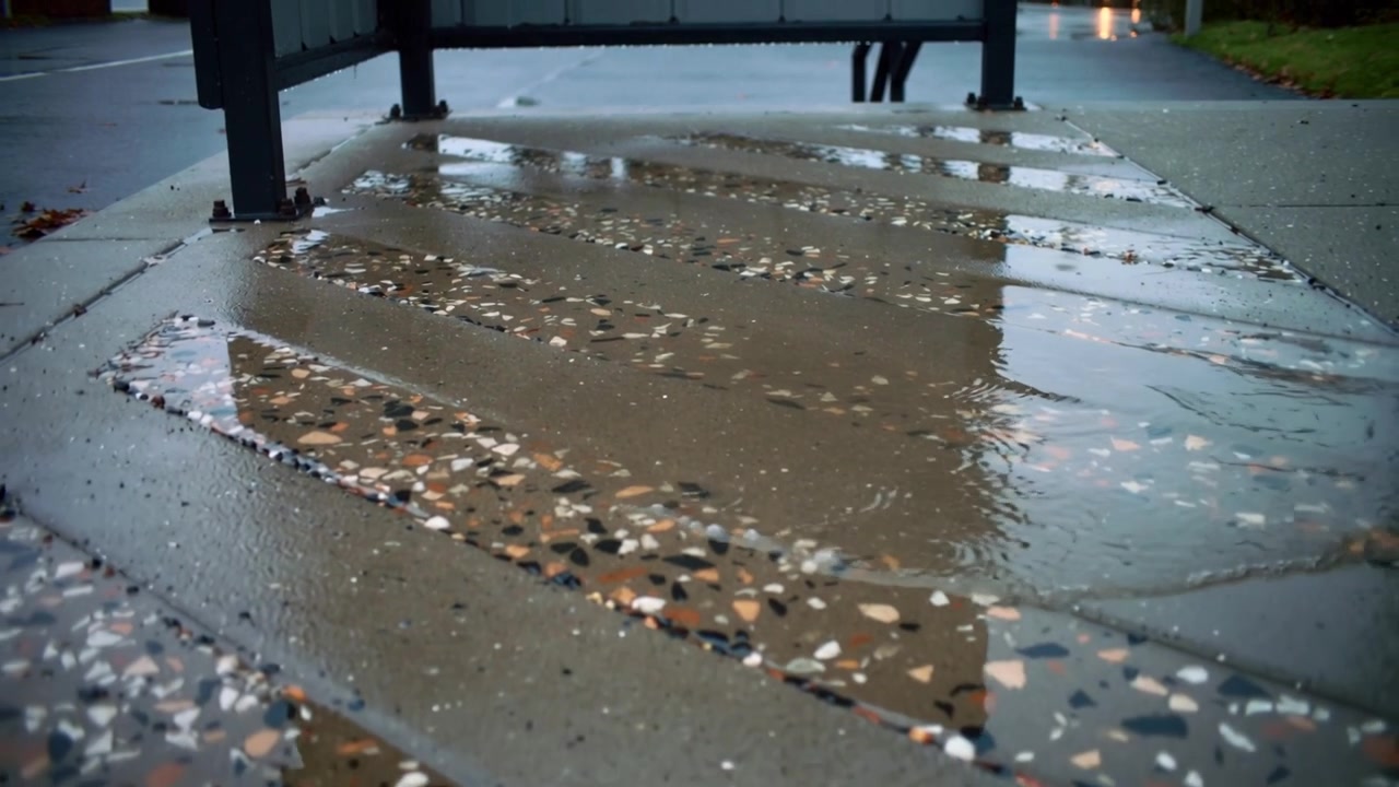 Creating ripples, light rain striking terrazzo panels beneath bench legs, water flowing toward curb