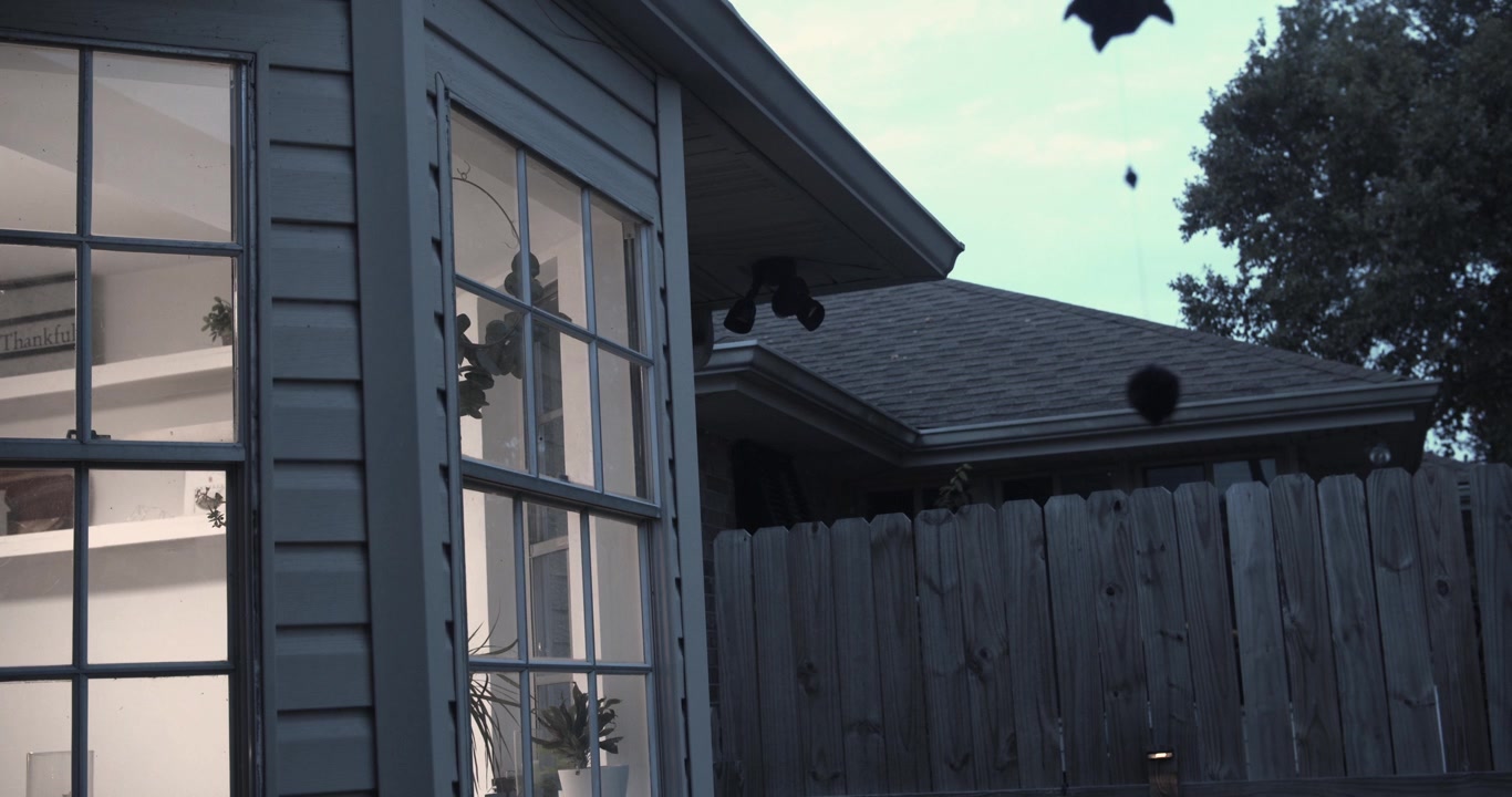 Cinematic view of suburban house bay windows exterior at dusk with dark leaves drifting across the frame, creating a moody autumn atmosphere and peaceful residential scene