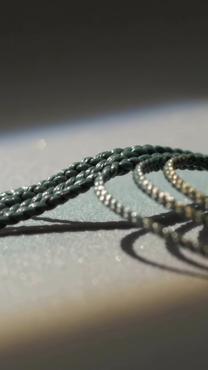Video verticale: La telecamera si sposta rivelando un cordino verde intrecciato su un tavolo, mostrando anelli di metallo e ombre.