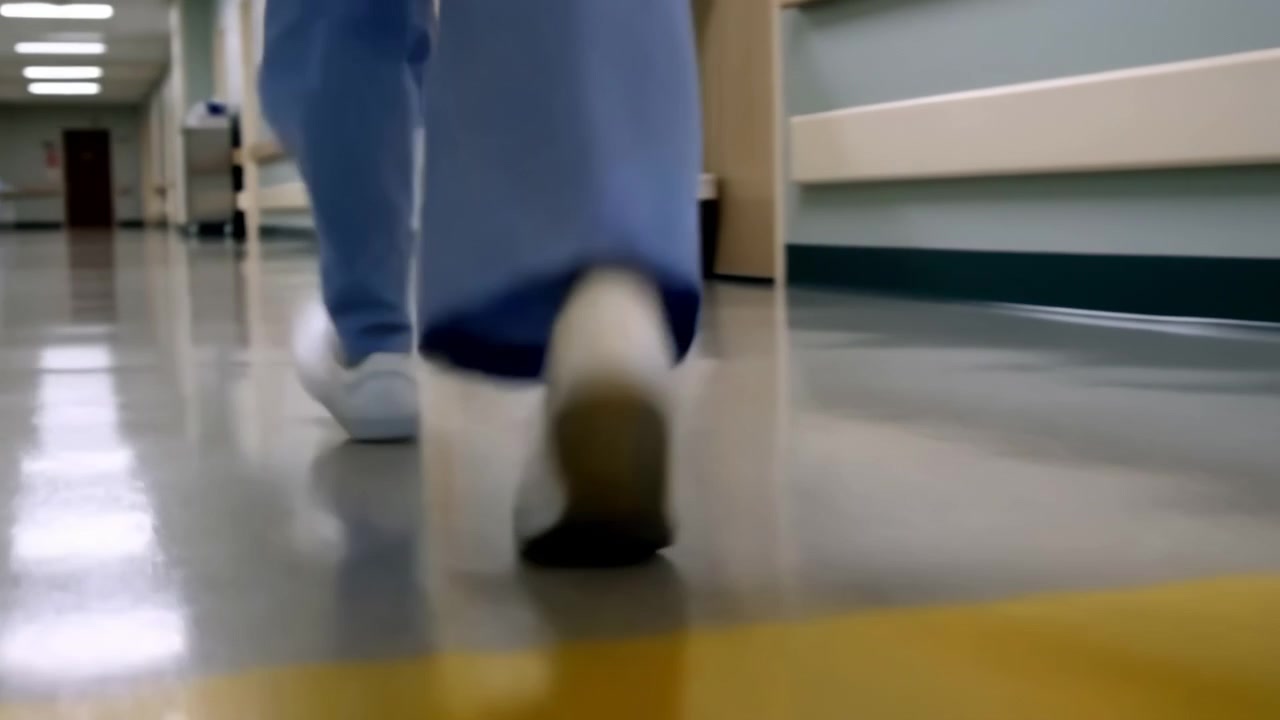 Close-up em ângulo baixo de um profissional de saúde de uniforme azul caminhando por um corredor de hospital polido, com portas e equipamentos desfocados e iluminação refletida no chão.