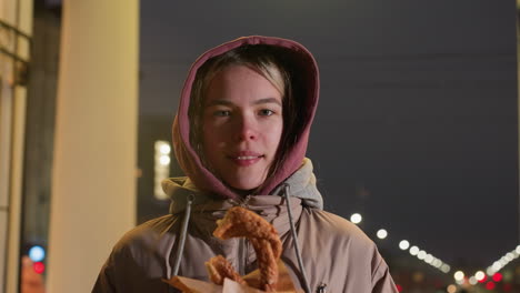 mujer joven sonriente de pie al aire libre por la noche sosteniendo pastel crujiente en la mano, brillo cálido del reloj de la calle iluminado en el fondo