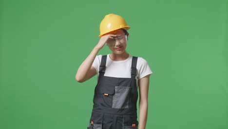 asian woman worker wearing goggles and safety helmet having a headache while standing in the green screen background studio