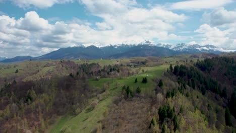 drone top view of a snow covered mountain with a clear blue sky, white clouds and green vegetation with a small house on the top of a hill, backwards movement, romania