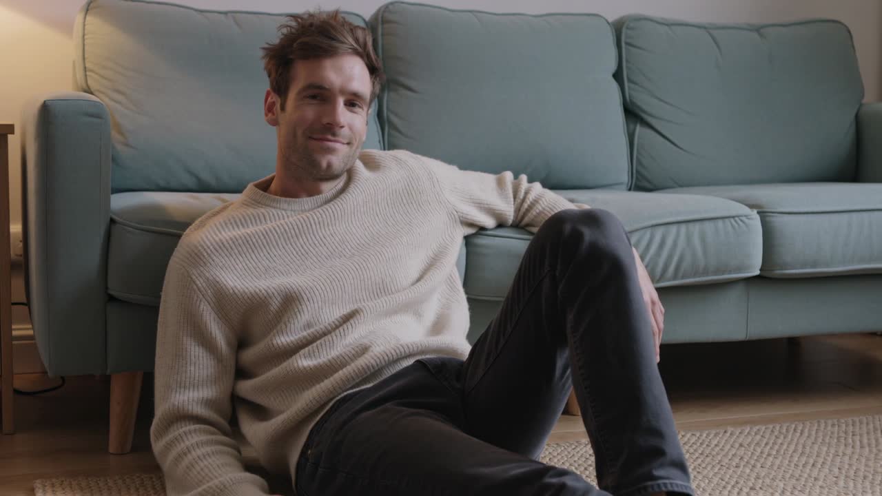 Telecamera che riprende un uomo adulto che si rilassa, sistemandosi nel maglione a casa, sorridendo e appoggiandosi a un divano color verde acqua.