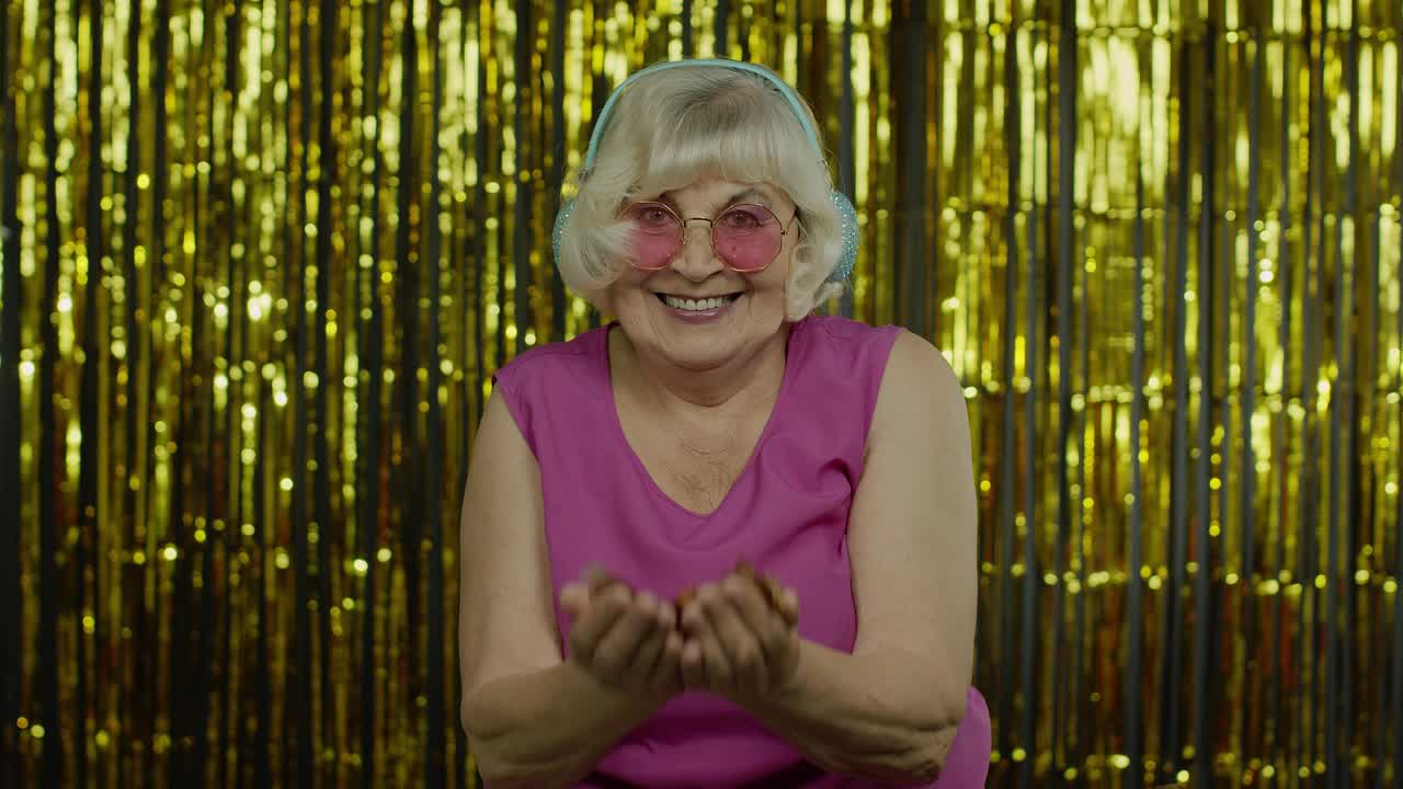 Felice donna anziana che ride, soffia coriandoli scintillanti, festeggia il compleanno, vince alla lotteria.