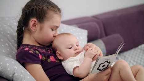 niña bonita leyendo un libro para niños con el bebé en casa. hermana sosteniendo al hermano bebé