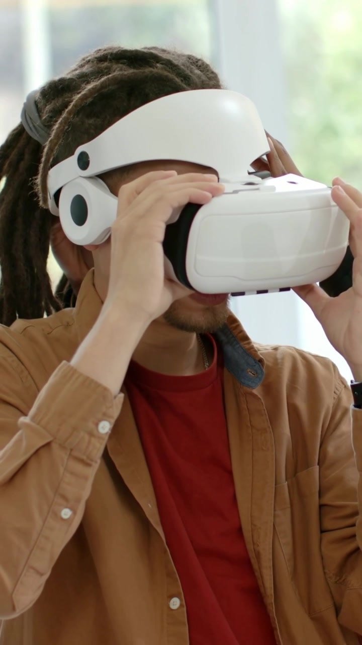 Video verticale: Uomo con camicia marrone che solleva un visore VR, regolando la cinghia a casa per un test, con l'aiuto di una mano.