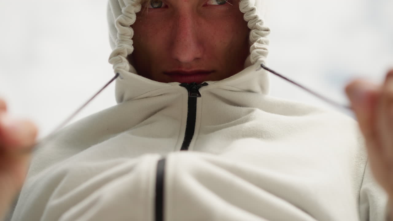 낮은 앵글에서 흰색 후드티를 조이고 있는 백인 남성, 흐린 하늘 배경, 위쪽 카메라 앵글, 단호한 표정, 조임끈을 꽉 조임, 준비와 결의에 찬 모습, 달리기 전 야외 의식