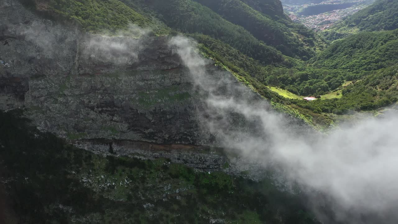 pared de acantilado épica y dramática con nubes delgadas que se mueven con algunas casas pequeñas detrás junto a campos de hierba verde y un bosque frondoso