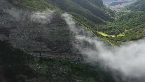 Epic-and-dramatic-cliff-wall-with-thin-clouds-moving-around-with-a-few-small-houses-behind-next-to-green-grass-fields-and-lush-forest