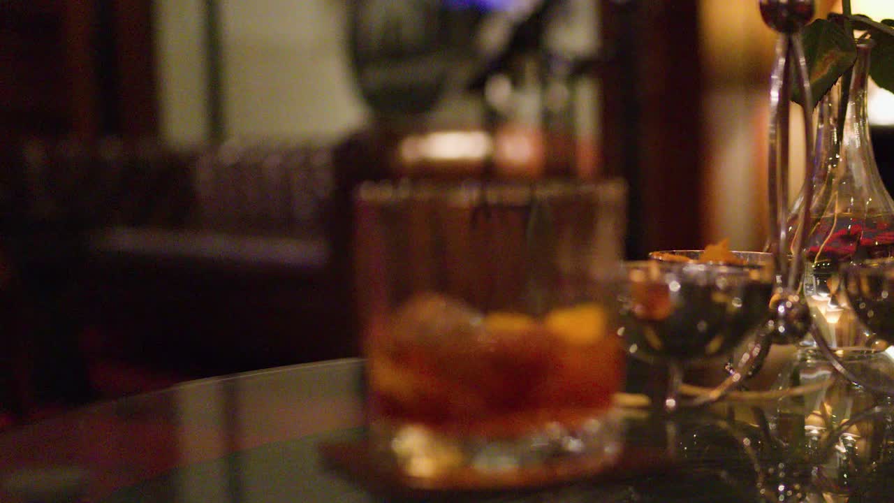 Whiskey glass with ice on reflective bar table, warm lounge lighting, slow focus shift