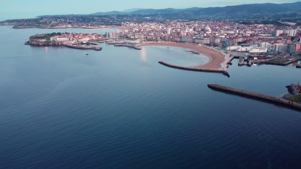 Riprese aeree panoramiche con drone del paesaggio urbano di Gijon al tramonto, con spiaggia, porto commerciale e turistico, nel nord della Spagna.