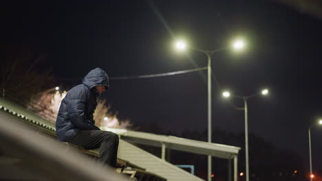 a man wearing a hood is seated alone on bleachers at night, with streetlights illuminating the surrounding area