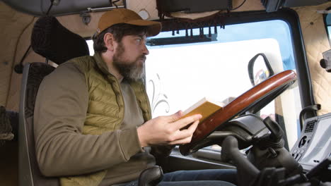 Side-View-Of-Worker-Sitting-In-A-Truck-In-A-Logistics-Park-While-Reading-A-Book