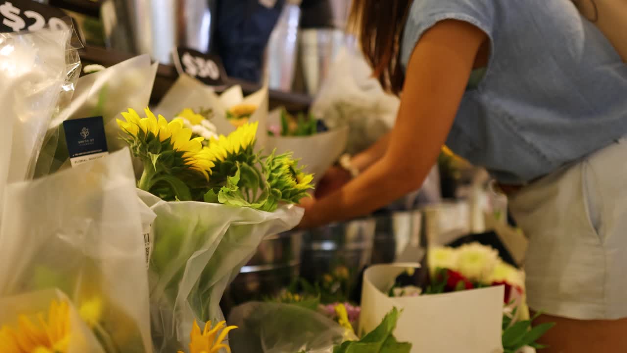 A woman in a blue shirt and white shorts selects flowers in a well-lit florist shop