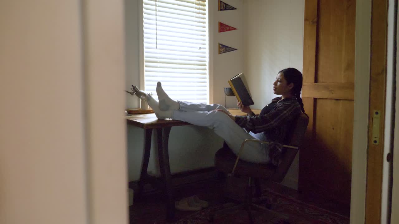 Relaxed Puerto Rican female reading captivating story book with feet raised on bedroom desk