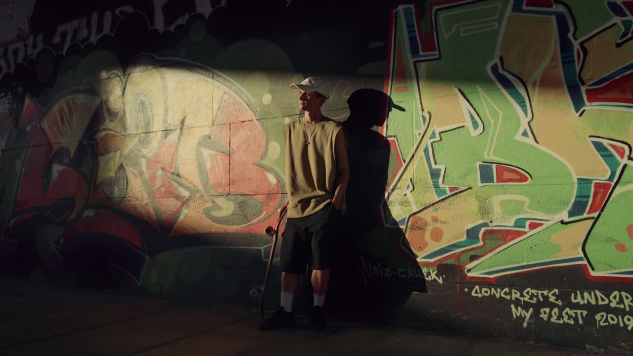 그래피티 벽 근처의  ⁇ 빛에서 스케이트 보드와 함께 휴식을 취하는 젊은  ⁇ 스터.