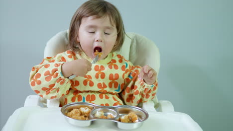 little girl eating tasty golden crispy chicken breast nuggets sitting in baby chair at home