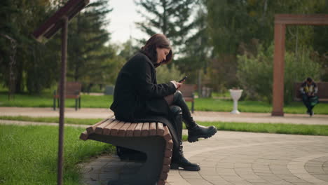 woman sitting on park bench using smartphone, dressed in black coat and boots, surrounded by lush green trees, relaxed outdoor atmosphere in urban park, blurred view of person on swing chair